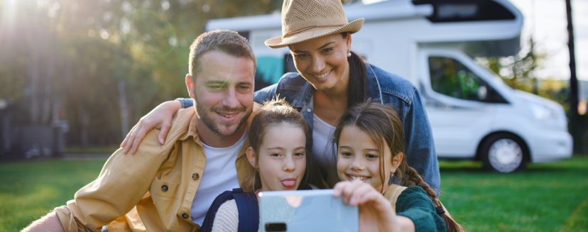 A happy young family with two children ltaking selfie with caravan at background outdoors.